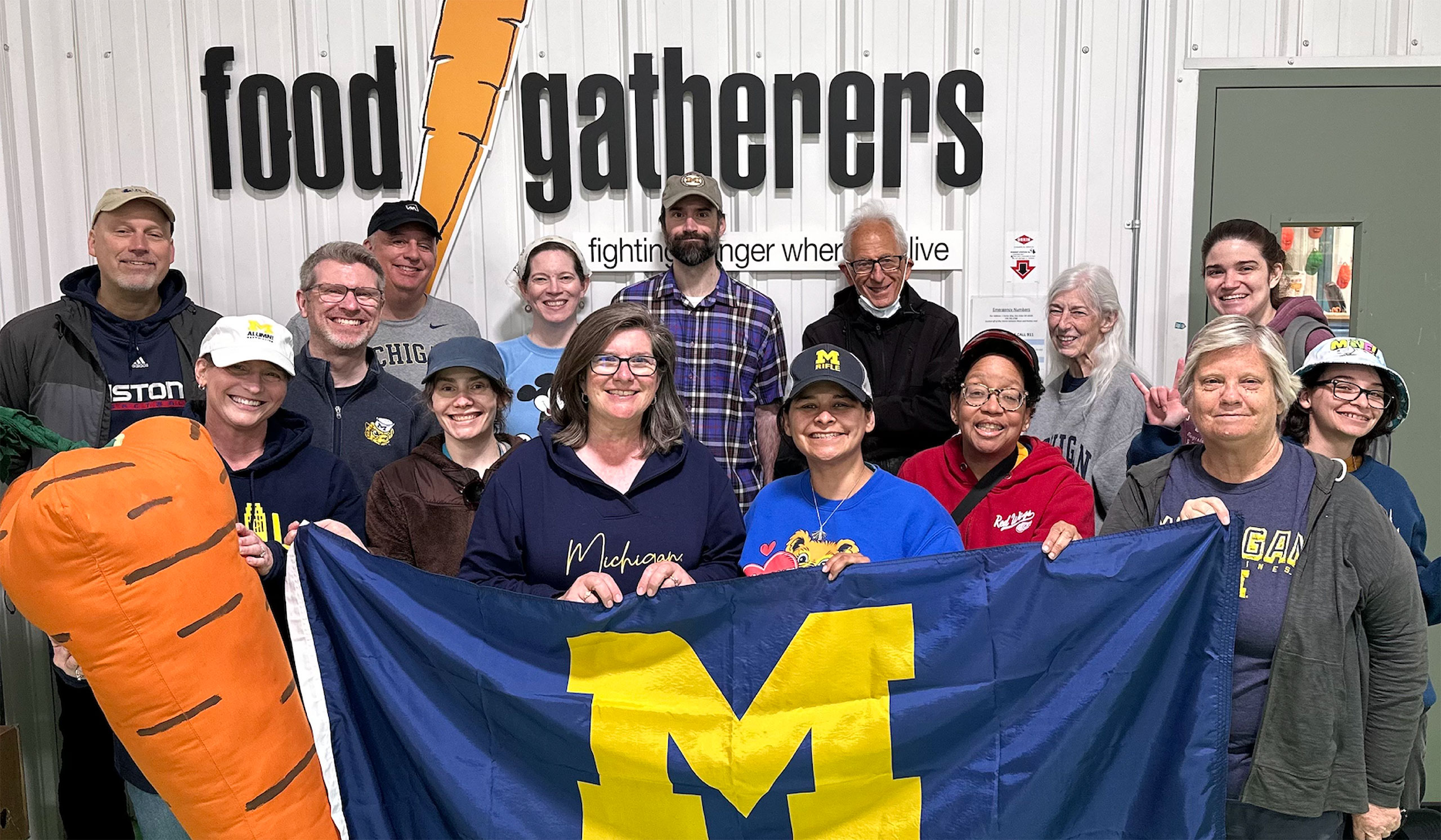 A group of smiling people pose indoors, holding a Michigan flag and a carrot prop in front of a Food Gatherers sign.
