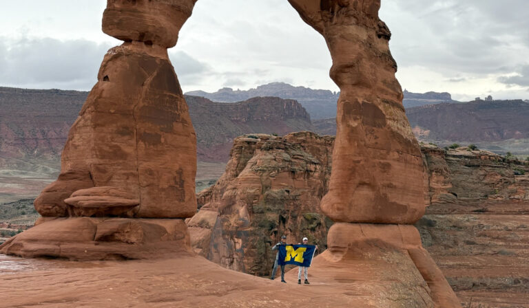 Two people under a sandstone arch in a rocky desert hold a blue flag with a yellow M, with clouds and mesas in the distance.