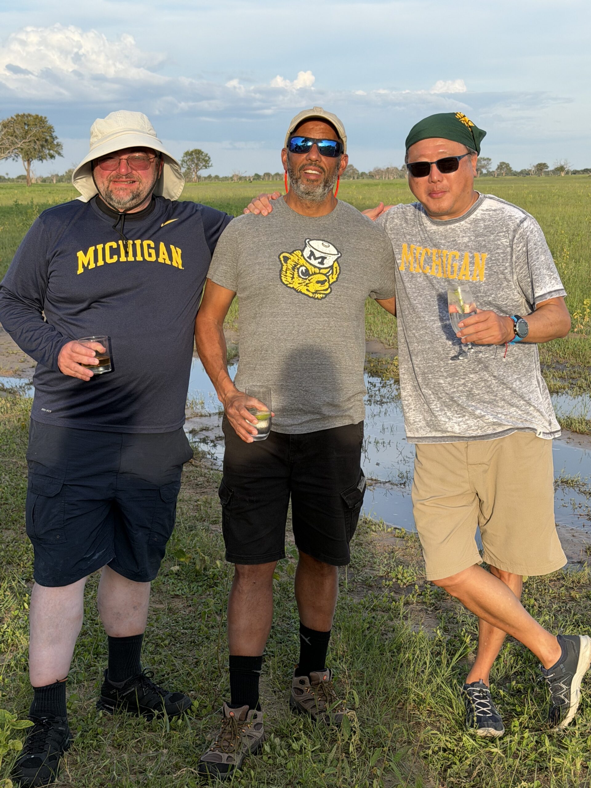 Three men wearing Michigan sports gear stand on a grassy field under the blue sky, smiling and holding drinks.