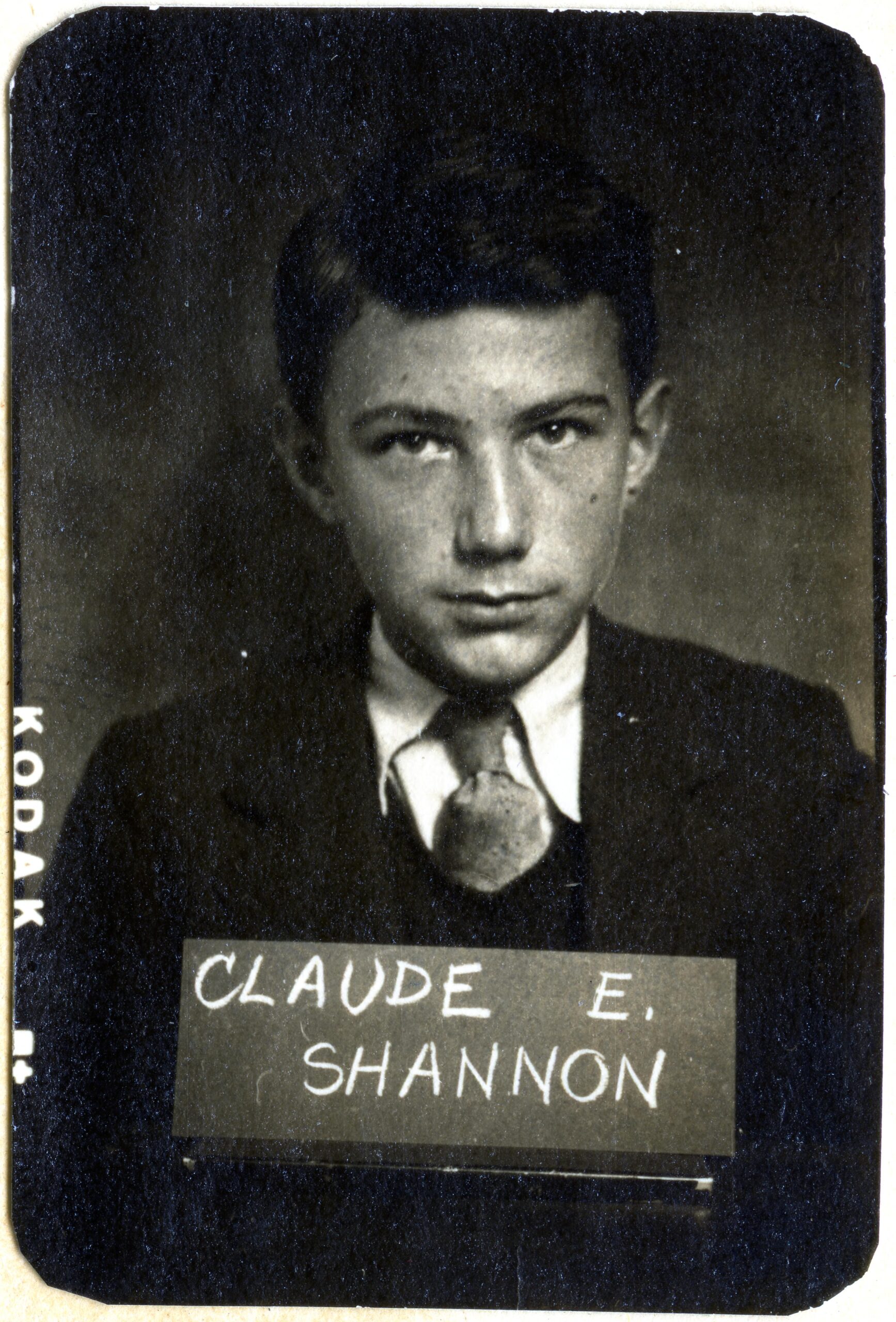 A student in a suit and tie poses with a hand written chalk sign that says "Claude E. Shannon"