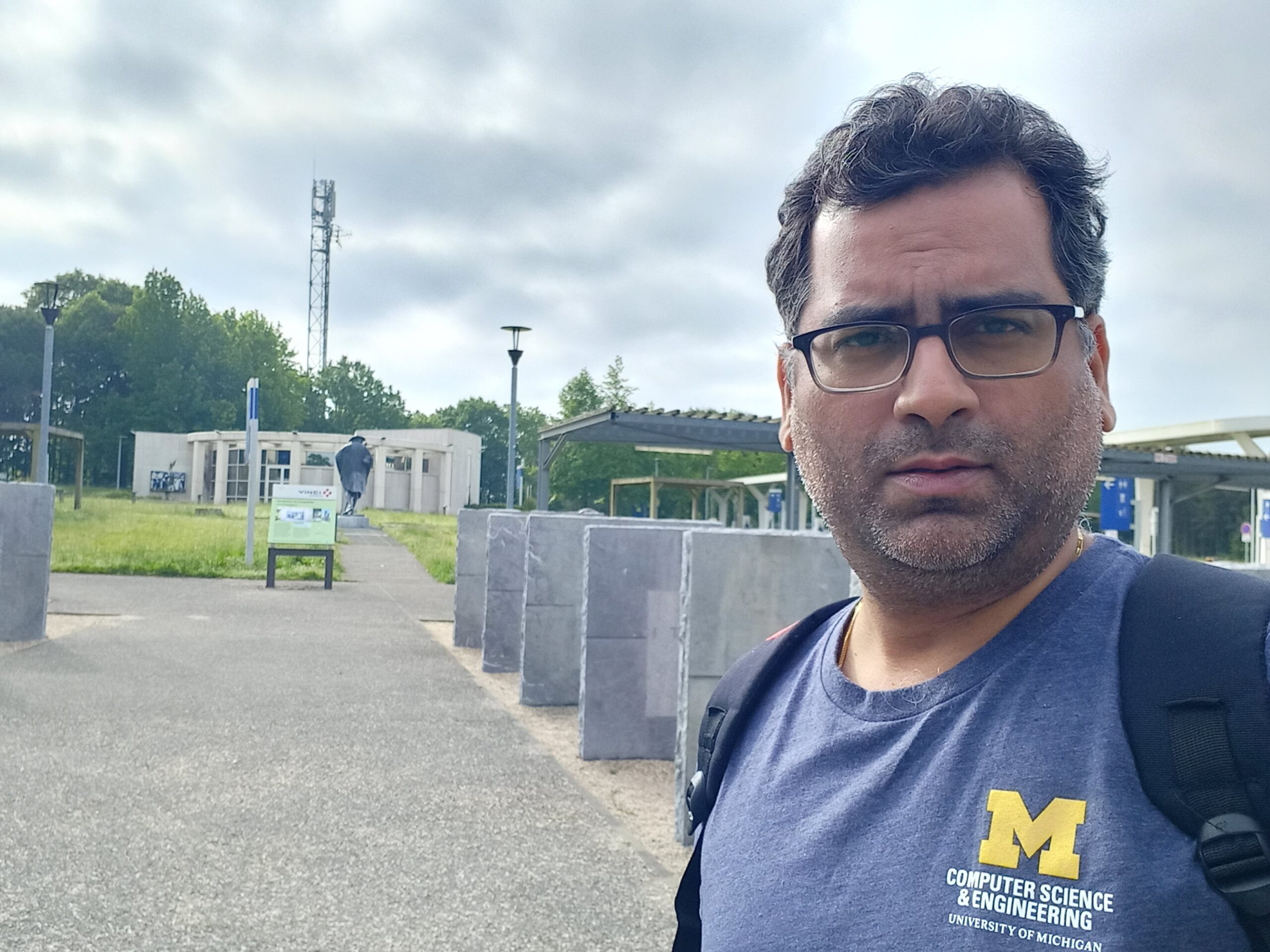 A man in glasses and a blue University of Michigan Computer Science & Engineering shirt stands outside with concrete pillars, greenery, and modern buildings behind him under a cloudy sky.