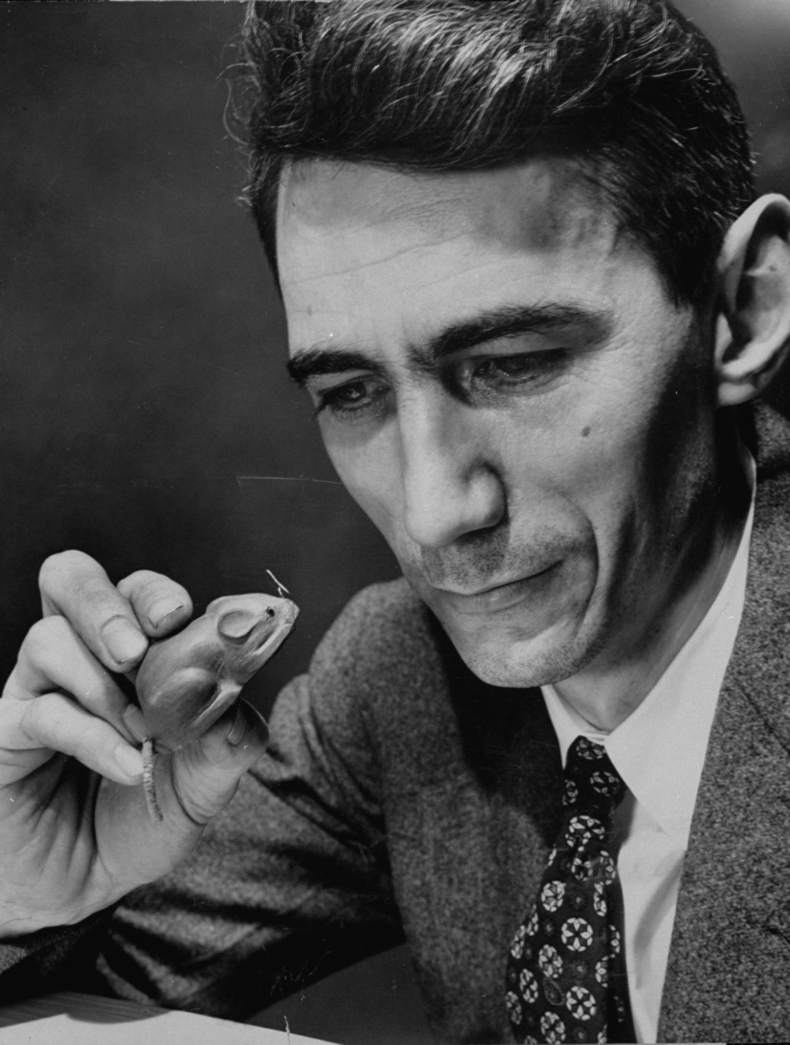 A man in a suit and patterned tie looks closely at a small mechanical mouse, appearing thoughtful, focused, and analytical.