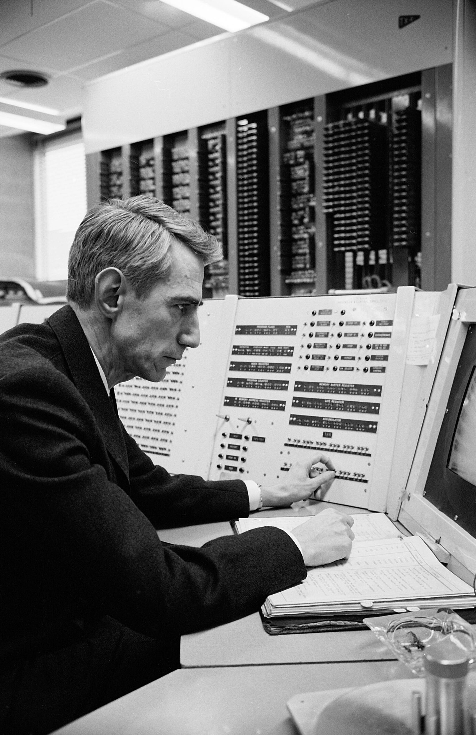 A man in a suit sits at a vintage control panel with buttons, switches, and an open notebook in a room of large computer equipment.
