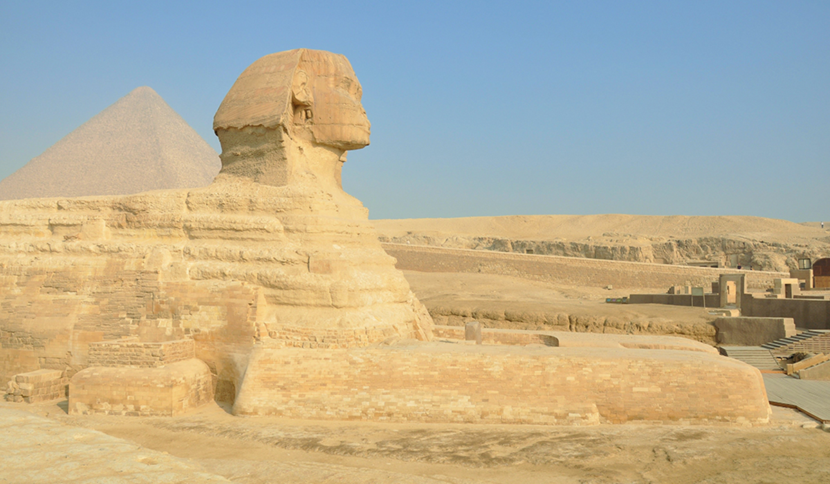 The Great Sphinx of Giza in Egypt shown in profile, with a pyramid rising in the background under a clear blue sky.