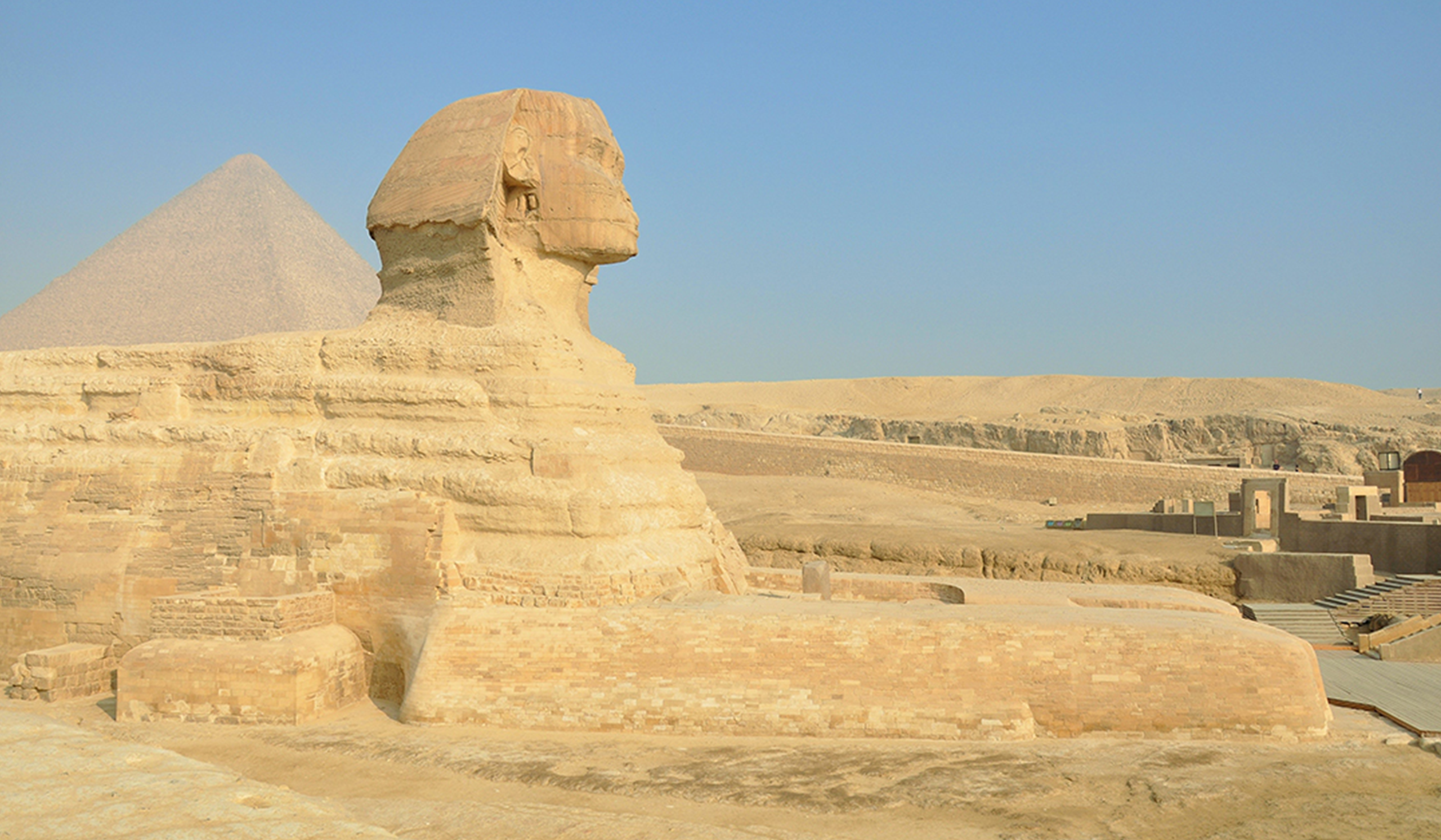 The Great Sphinx of Giza in Egypt shown in profile, with a pyramid rising in the background under a clear blue sky.