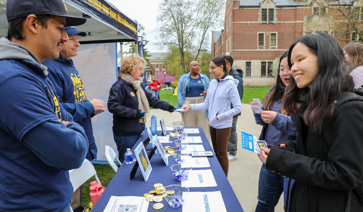 Students interact with staff at an outdoor campus event table, picking up brochures and buttons. A blue tablecloth displays materials, and people are smiling and talking. Buildings and trees are visible in the background.