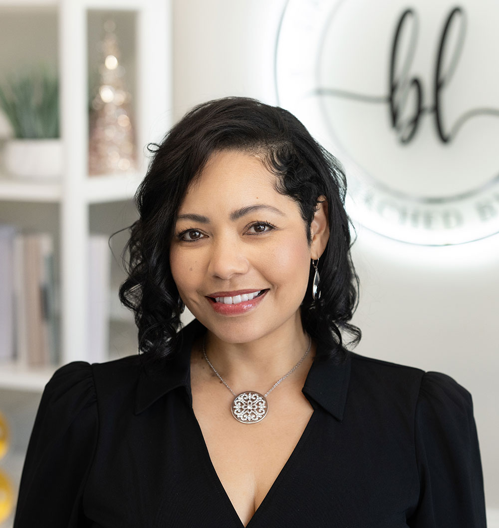 A smiling woman with wavy dark hair wearing a black top and a silver necklace stands in a modern, light-filled office, with shelves and a decorative sign in the background.