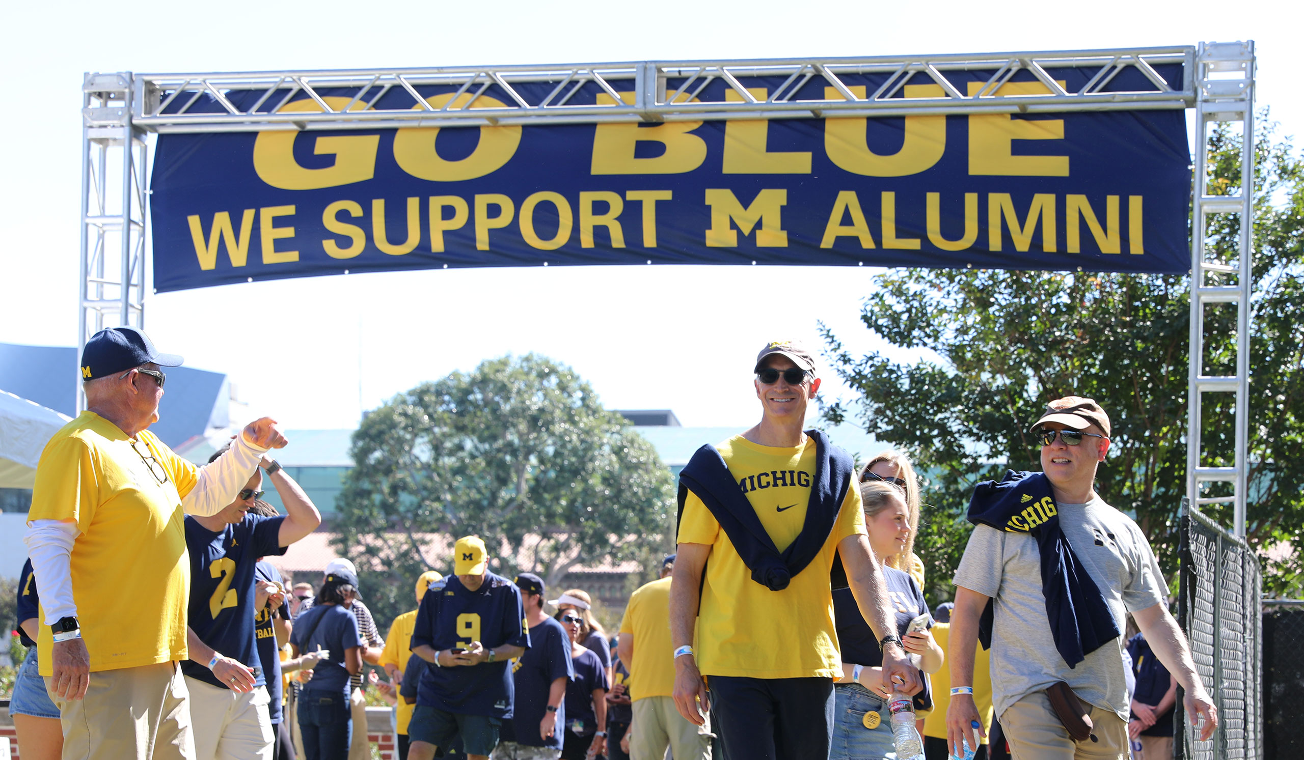 Fans in Michigan apparel enter a tailgate, walking beneath a banner that says GO BLUE WE SUPPORT M ALUMNI.