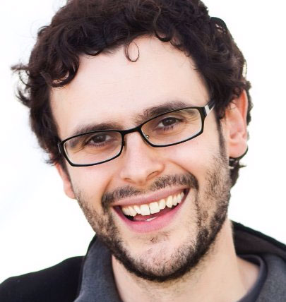 A man with curly brown hair, a beard, and glasses smiles at the camera. He wears a dark jacket over a gray shirt, with a white background behind him.