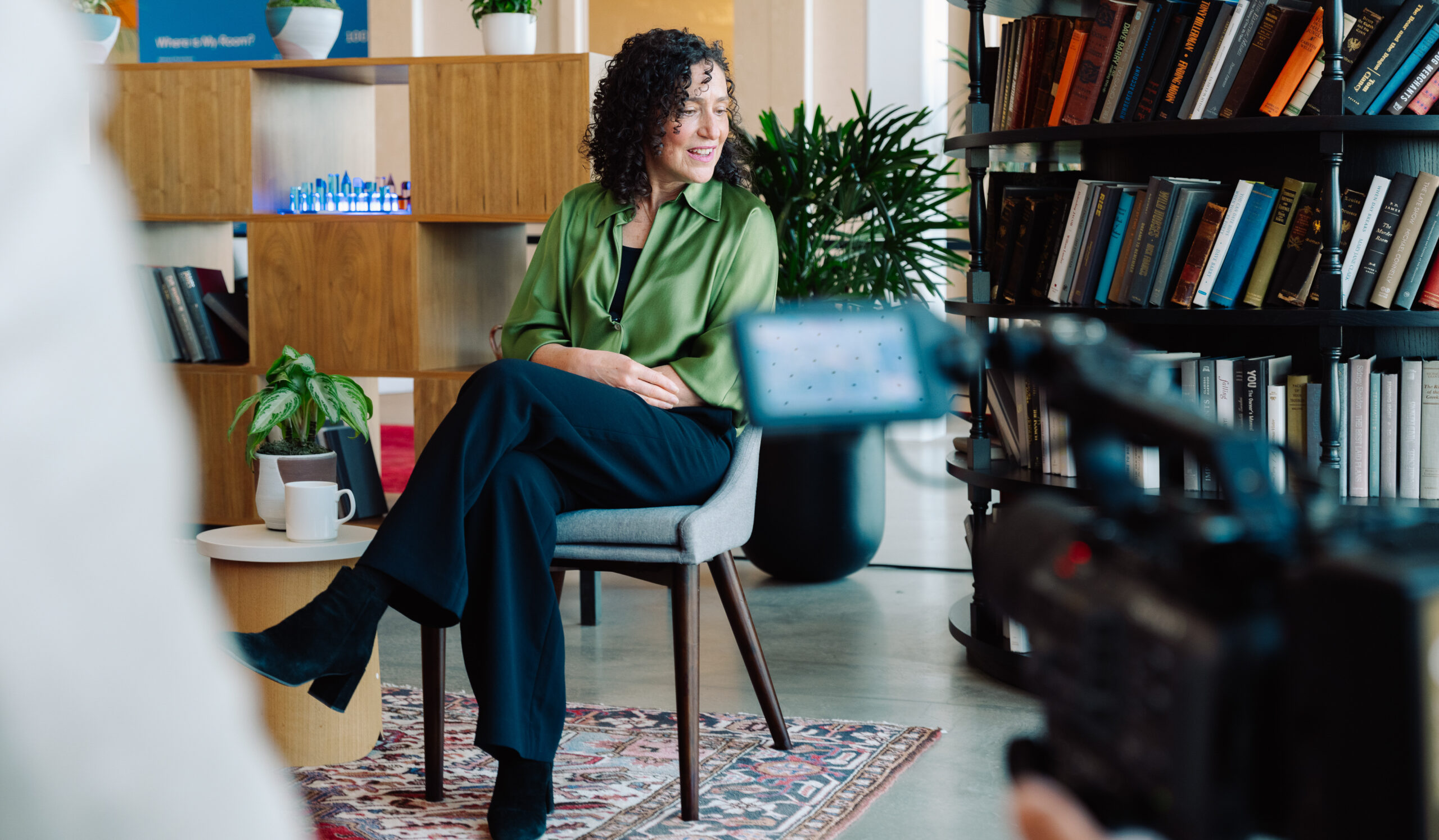 A woman with curly hair sits on a chair in a modern, cozy room with bookshelves, a table, and plants, smiling at a video camera.