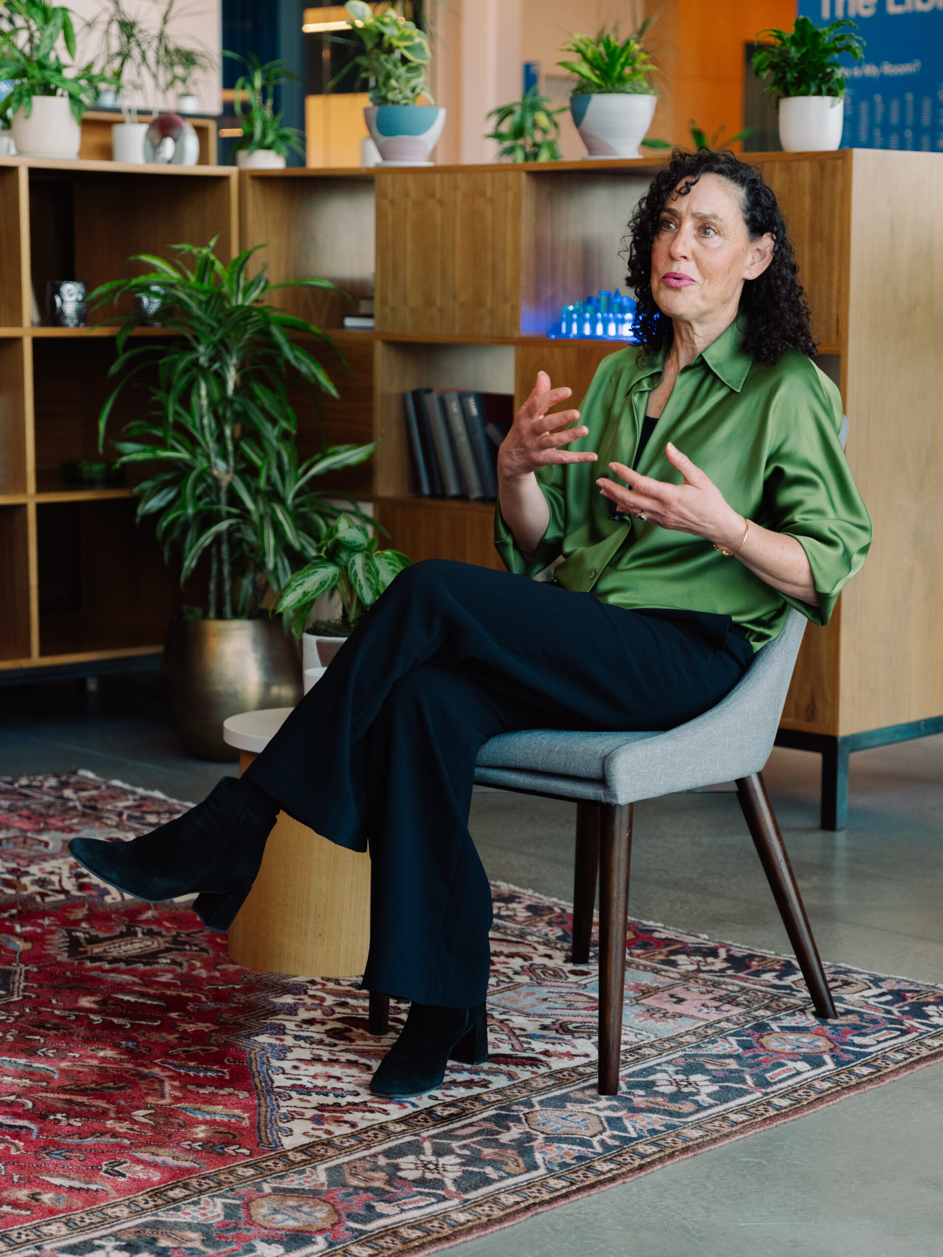 A woman with curly hair sits on a chair in a modern, cozy room with bookshelves, a table, and plants, speaking with her hands.