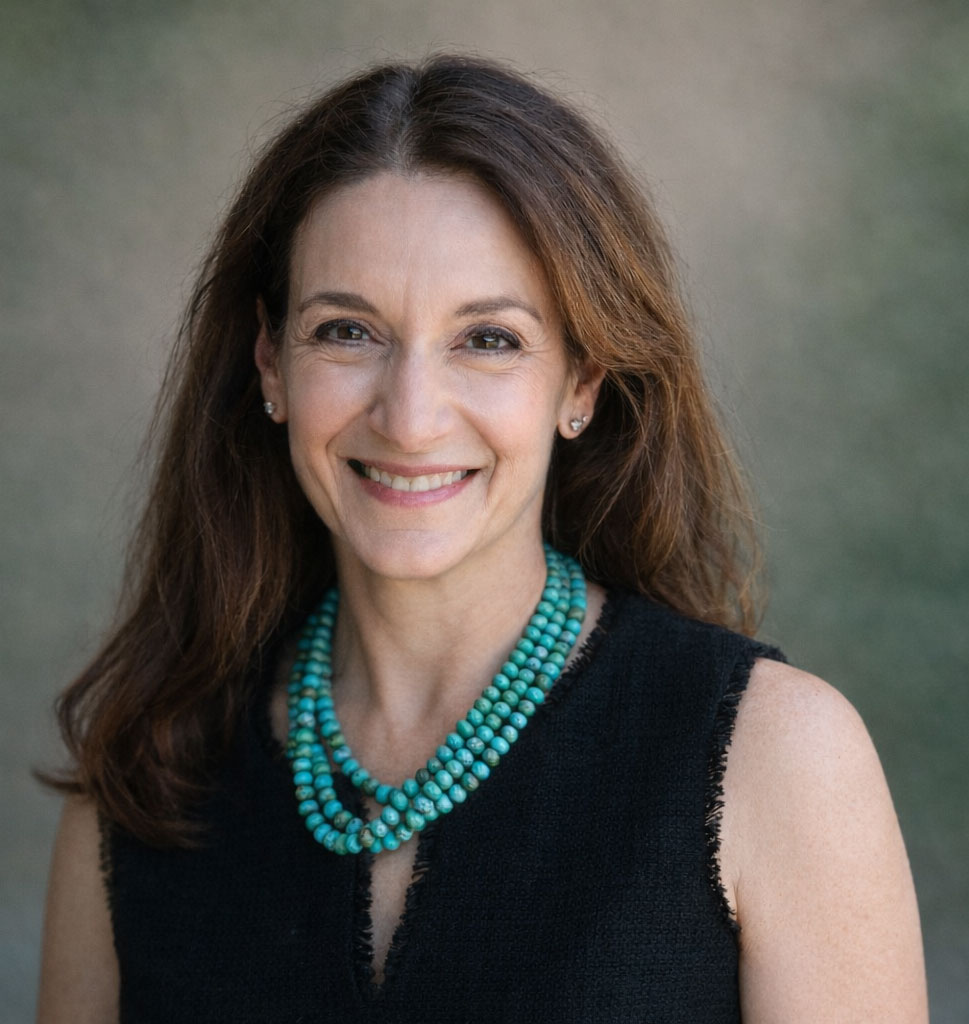 A woman with long brown hair smiles, wearing a sleeveless black top and a turquoise beaded necklace, in front of a blurred, neutral background.