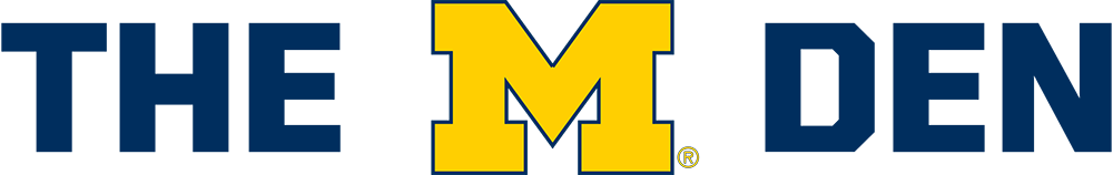 Logo featuring the words "THE M DEN," with "THE" and "DEN" in navy blue, separated by a large maize Block M.