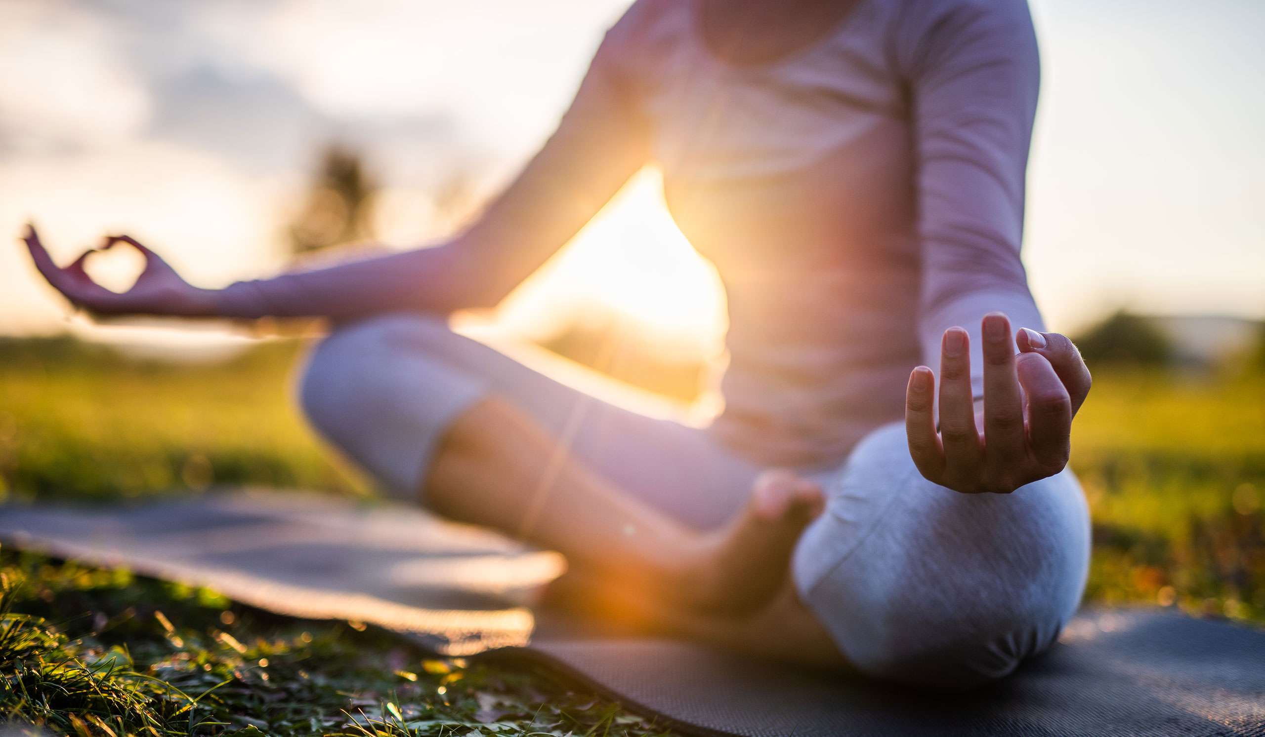 A person sits cross-legged on a yoga mat outdoors at sunset, wearing light-colored clothing, with hands resting on knees in a meditation pose.
