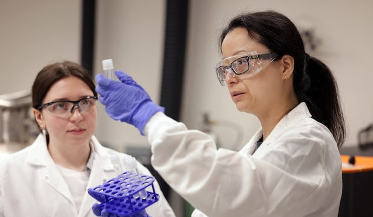 Two scientists in lab coats and goggles examine a test tube in a lab, with equipment and devices visible.