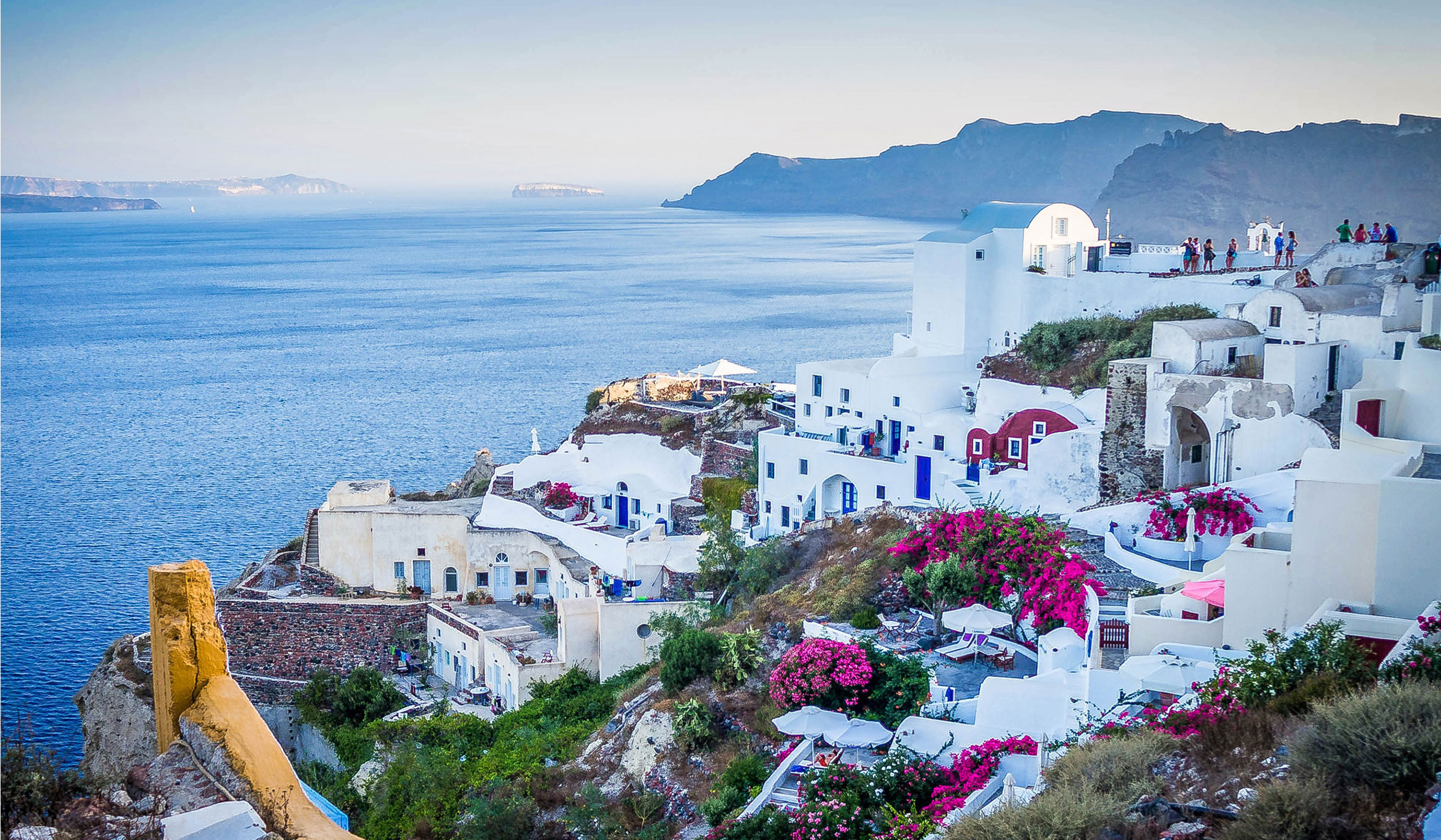 Whitewashed buildings sit atop a cliff overlooking a calm blue sea, with vibrant pink flowers and distant islands beneath a clear sky. Tourists enjoy the scenic view.