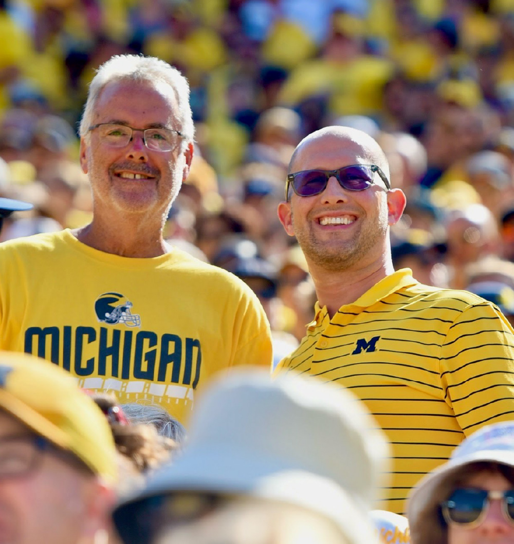 Two people wearing maize Michigan shirts smile at Michigan Stadium during a football game, surrounded by a crowd also dressed mostly in maize.