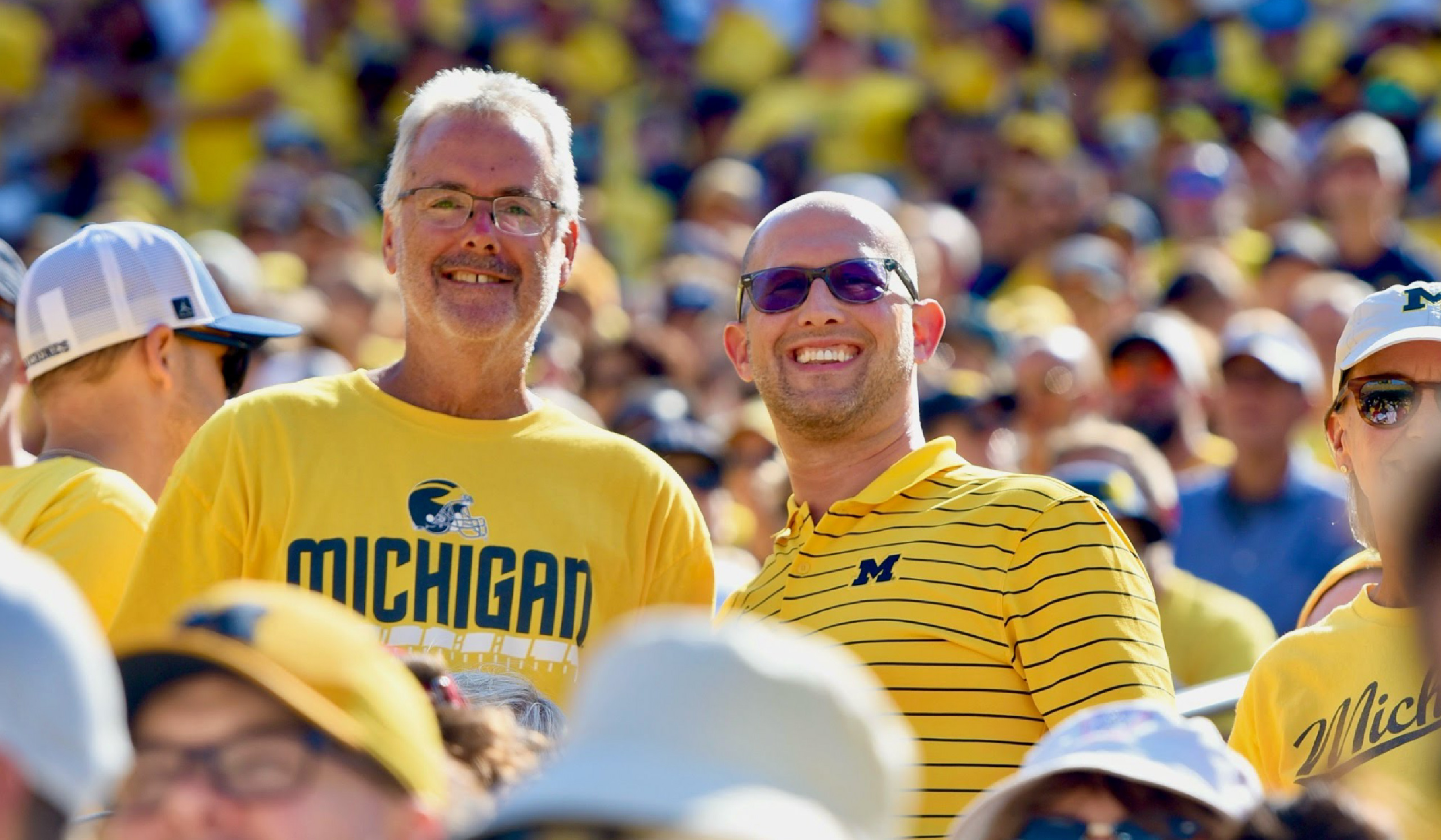 Two people wearing maize Michigan shirts smile at Michigan Stadium during a football game, surrounded by a crowd also dressed mostly in maize.