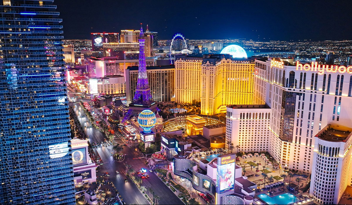 Aerial view of the Las Vegas Strip at night, featuring brightly lit hotels, casinos, neon signs, and replicas of famous landmarks such as the Eiffel Tower, with city lights visible in the background.