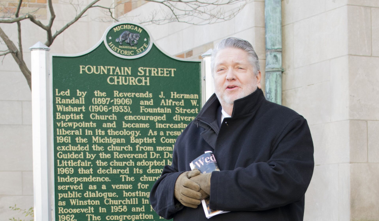 A person in a black coat stands in front of a historic marker for Fountain Street Church, holding a book.