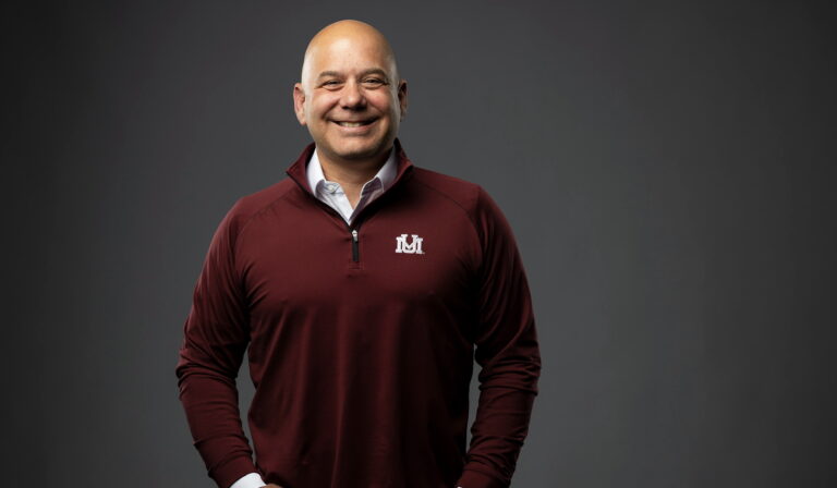 A person stands smiling, wearing a maroon quarter-zip pullover with a white logo and a collared shirt underneath.