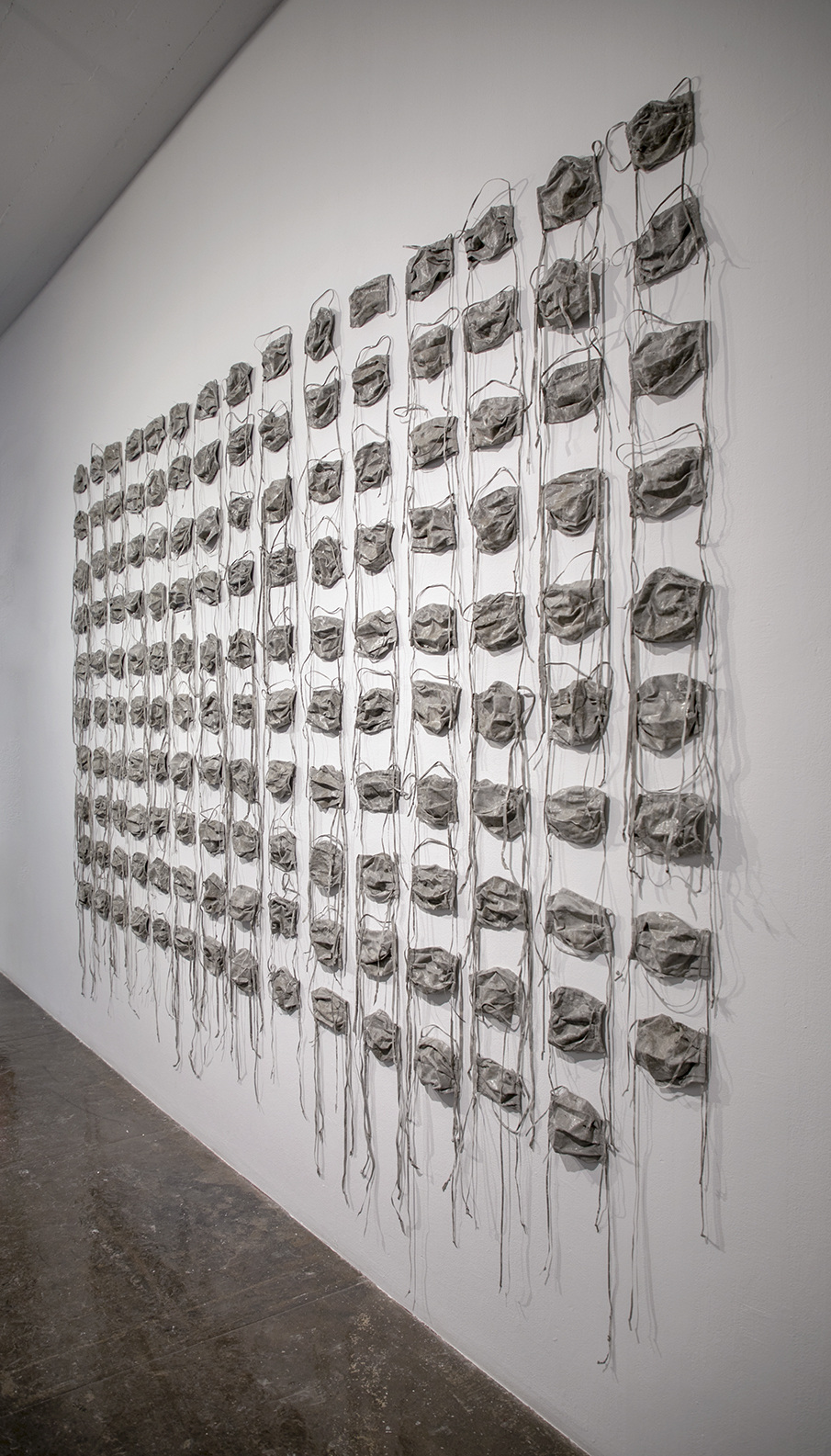 Disposable face masks dipped in grey concrete hang in rows on a white wall, each with loose strings dangling.