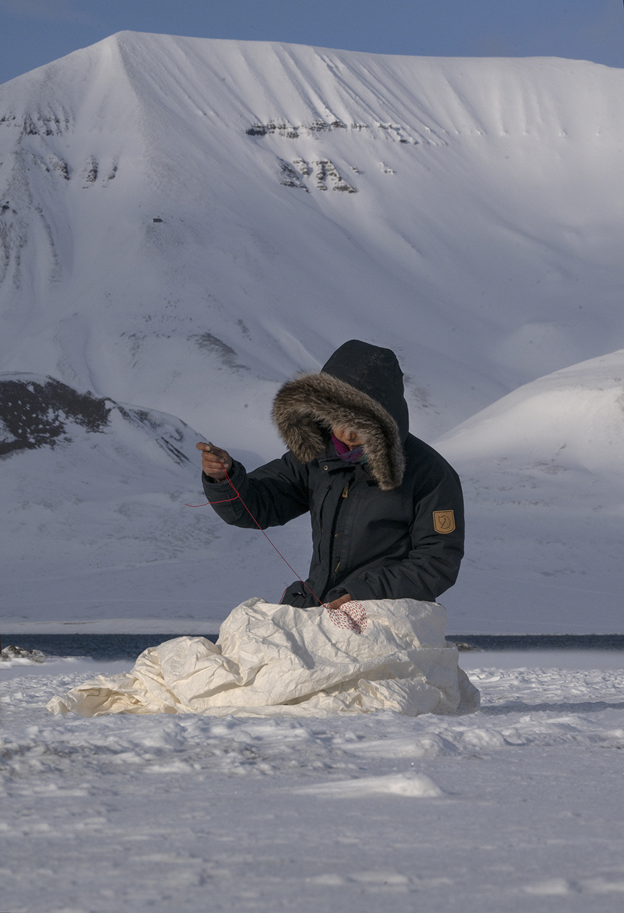Person in a parka with a fur-lined hood sits on snow, using string on white material, with snowy mountains in the background.