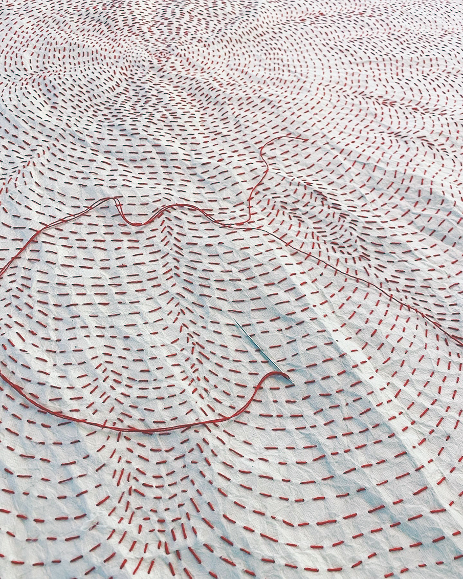A needle with red thread lies on white fabric decorated with swirling patterns of red dashed stitching.