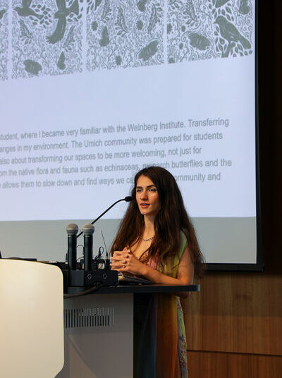 Mural design winner Suher Salim stands at a podium presenting a slide with intricate black-and-white artwork and descriptive text in a modern room.