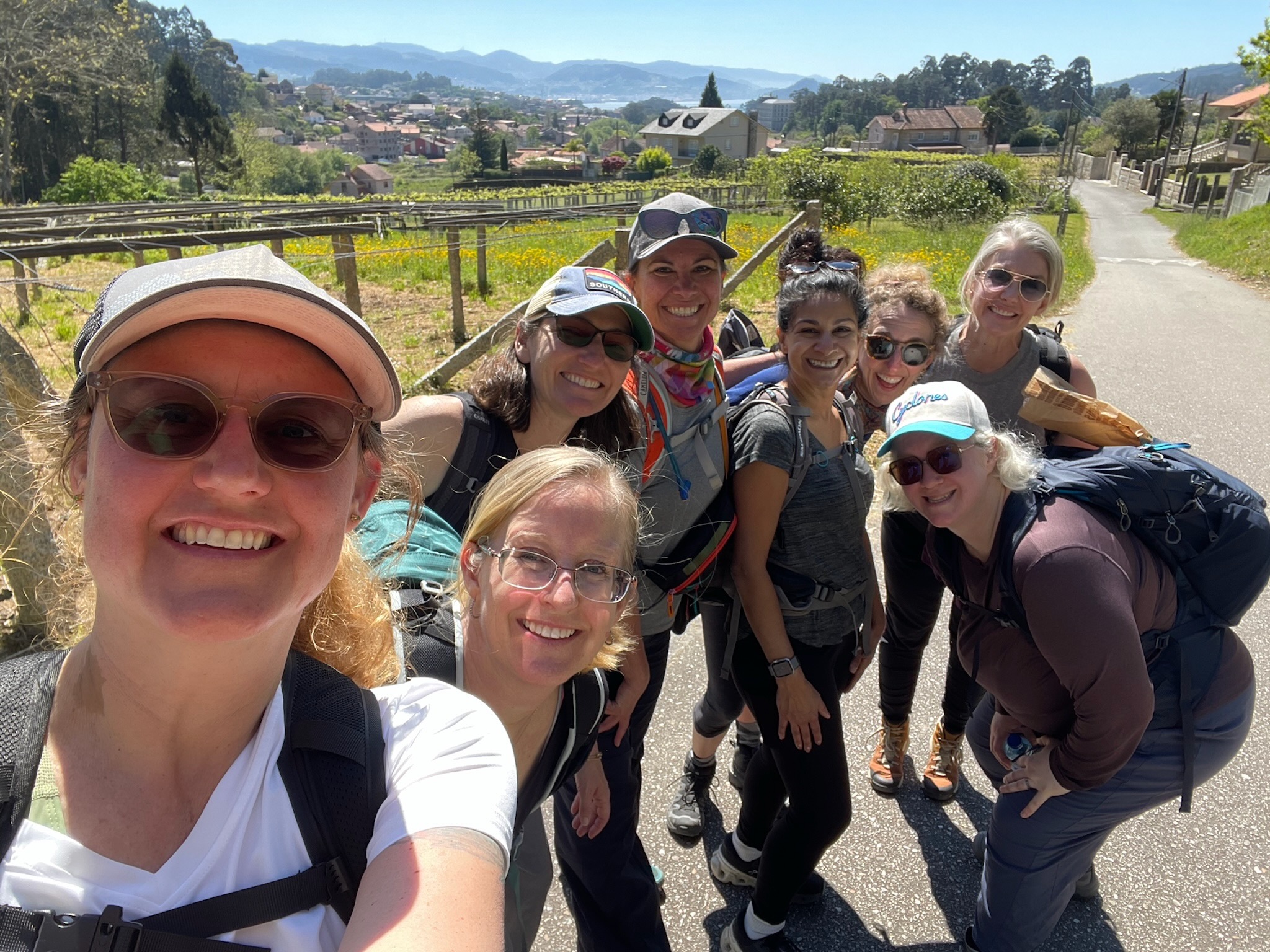 Eight smiling women wearing outdoor gear pose for a group selfie on a sunny trail, with green hills, houses, and mountains in the background, capturing the spirit of Michigan friendships forged during their Michigan journey.