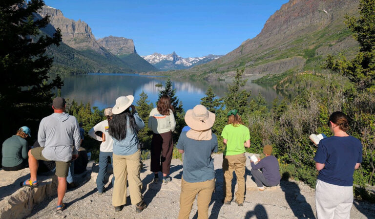 People stand and sit facing a mountain lake, surrounded by trees, under a clear blue sky.