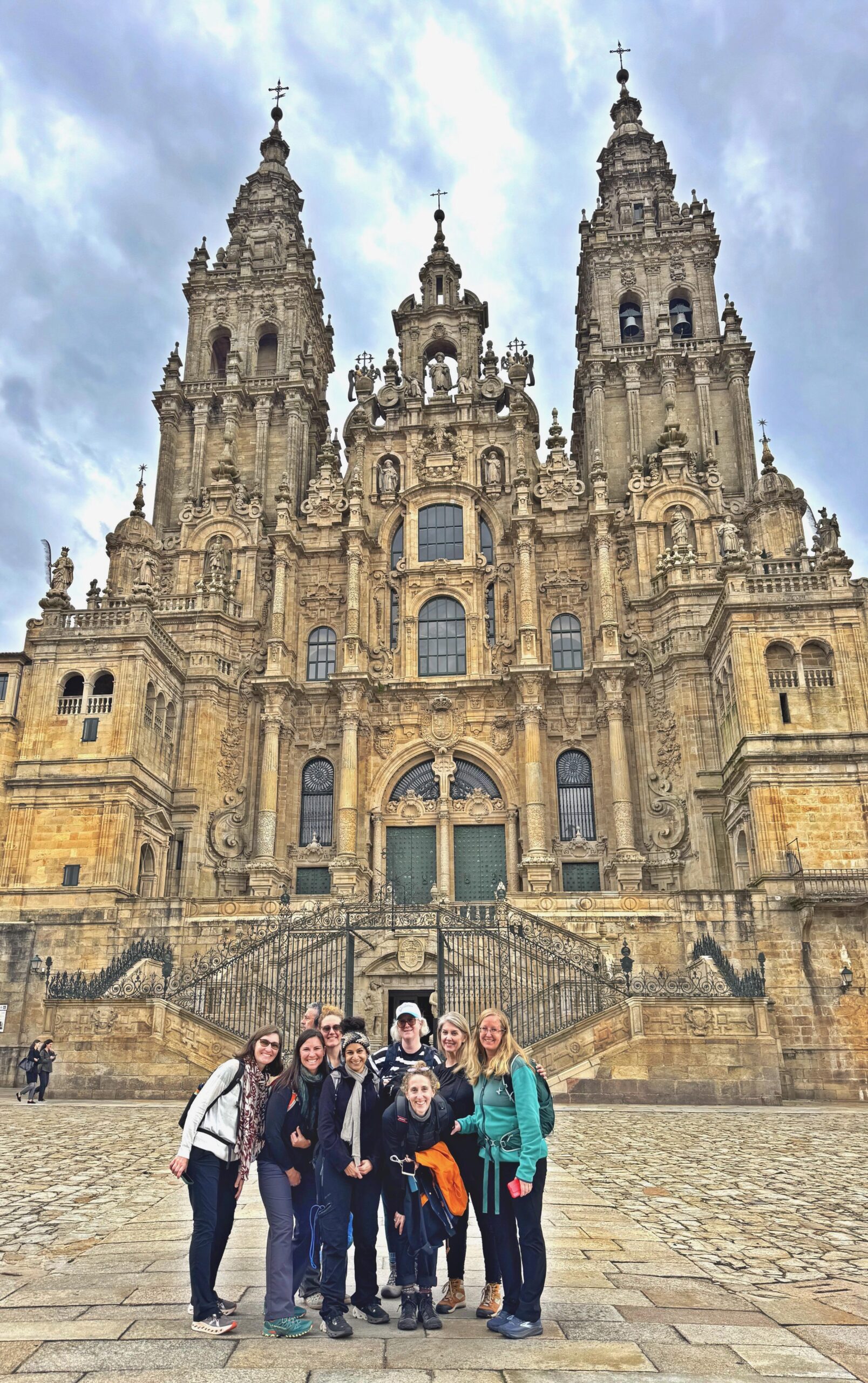 A group of people pose and smile in front of the ornate facade of the Cathedral of Santiago de Compostela, under a cloudy sky, their laughter reflecting how Michigan friendships can enrich the journey.