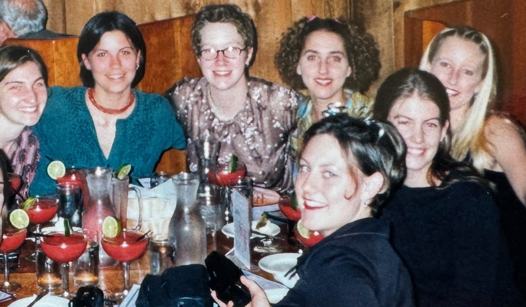 Seven women sit closely together at a restaurant table with several red cocktails, each topped with a lime. They smile at the camera, capturing a joyful moment during their Michigan journey. Wooden paneling and booth seats are visible in the background.