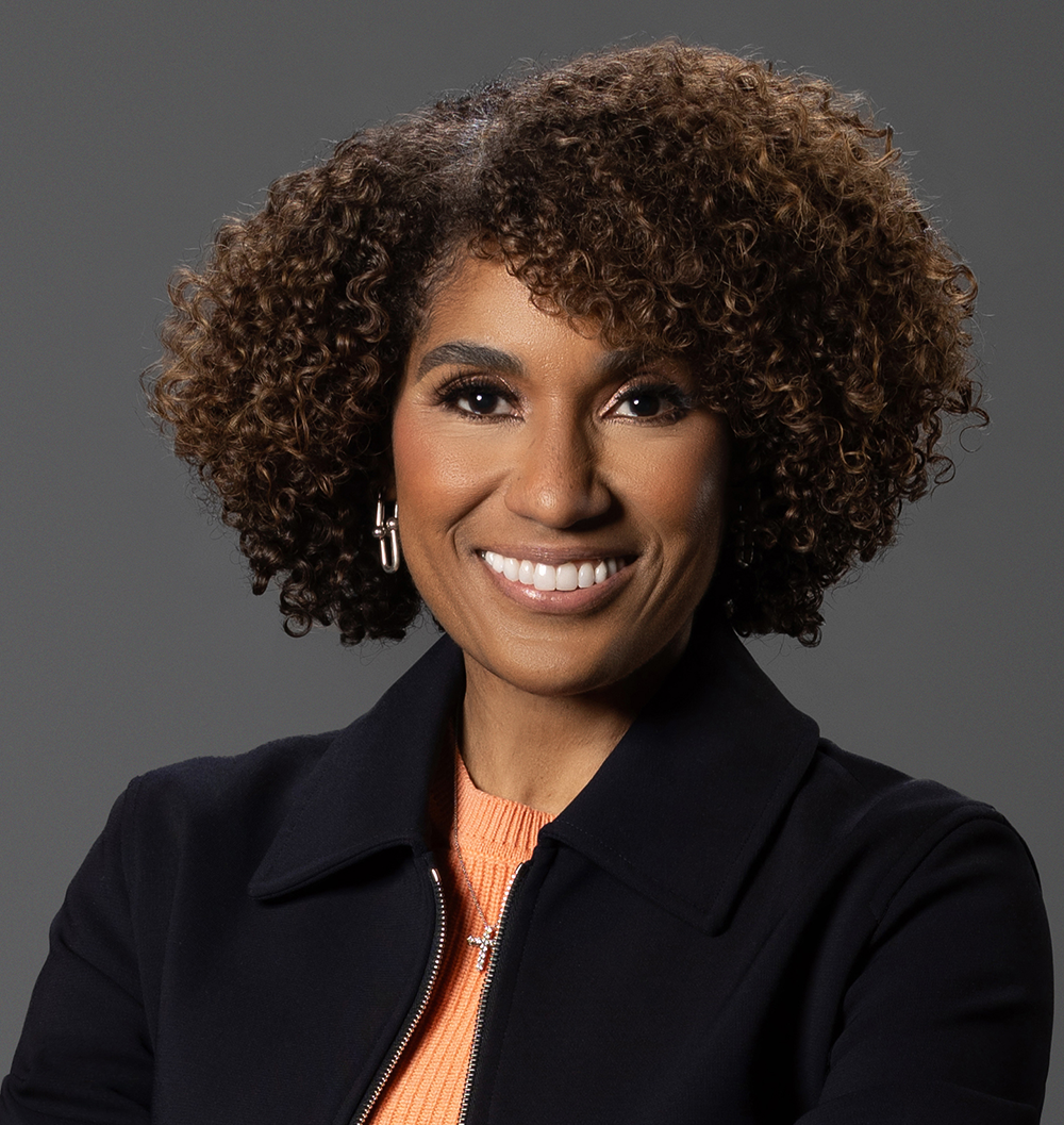 Smiling person with curly brown hair in a dark jacket and orange top, standing against a gray background.