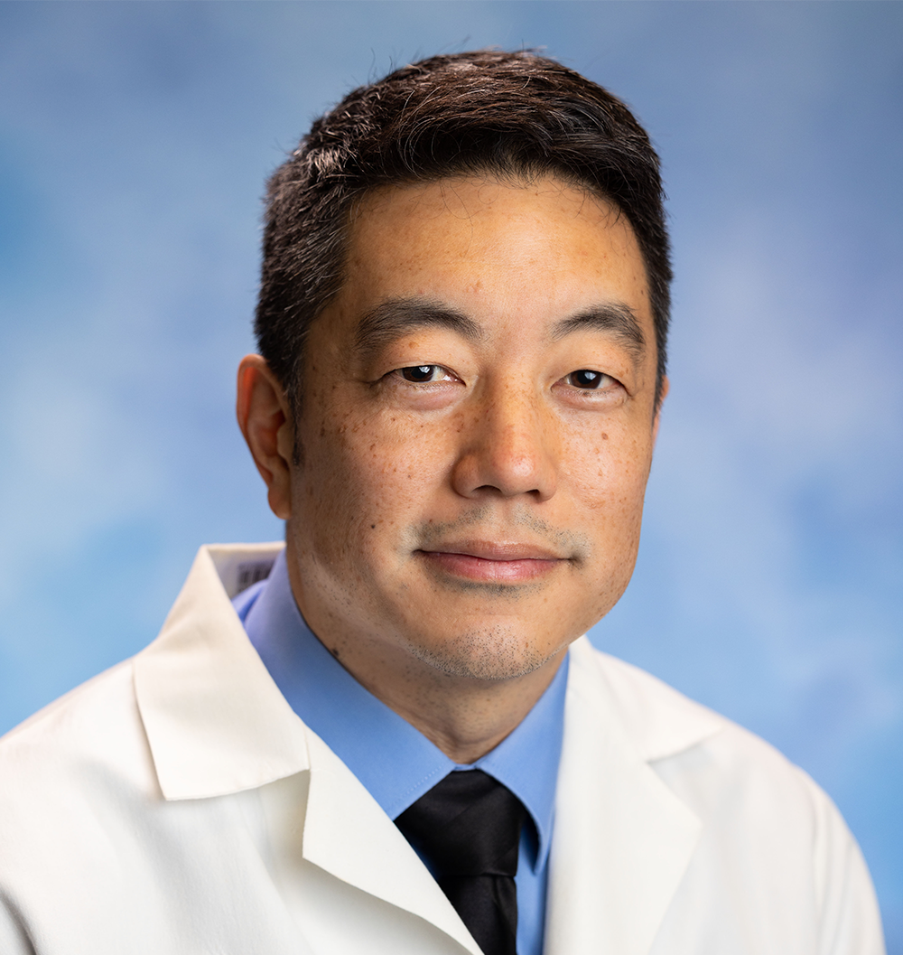 Person wearing a white lab coat, blue shirt, and black tie, posing against a light blue background.