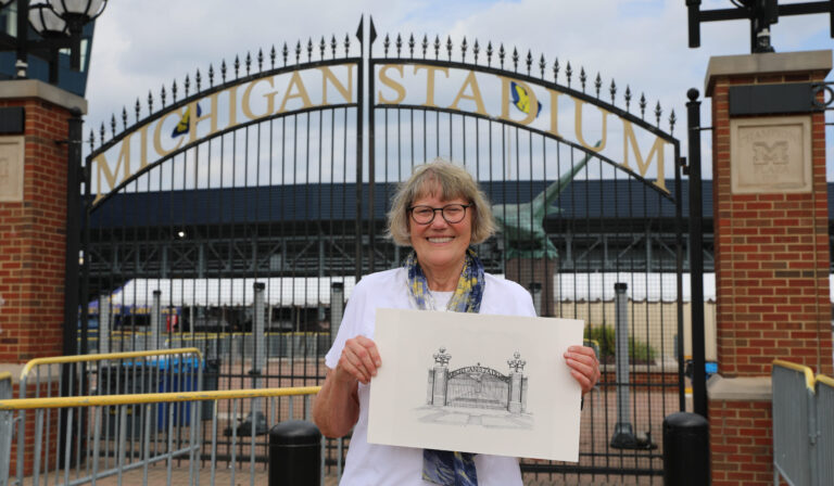 Smiling Judy Renfrew Hart holds a drawing of the stadium gate in front of Michigan Stadium’s entrance gate on a cloudy day.