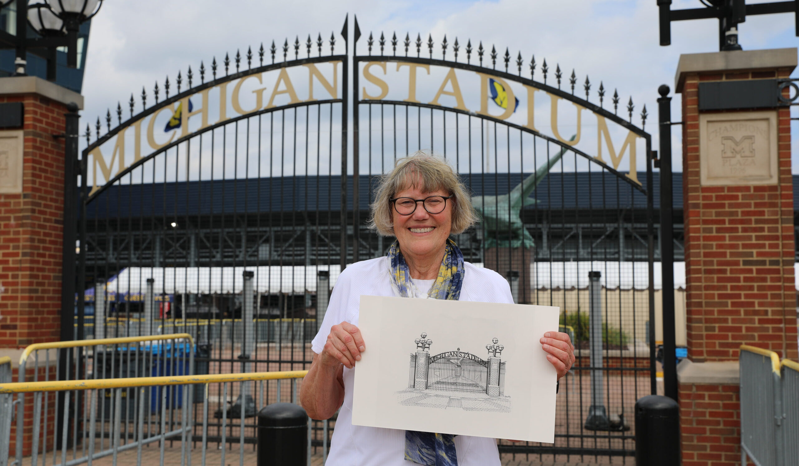 Smiling Judy Renfrew Hart holds a drawing of the stadium gate in front of Michigan Stadium’s entrance gate on a cloudy day.