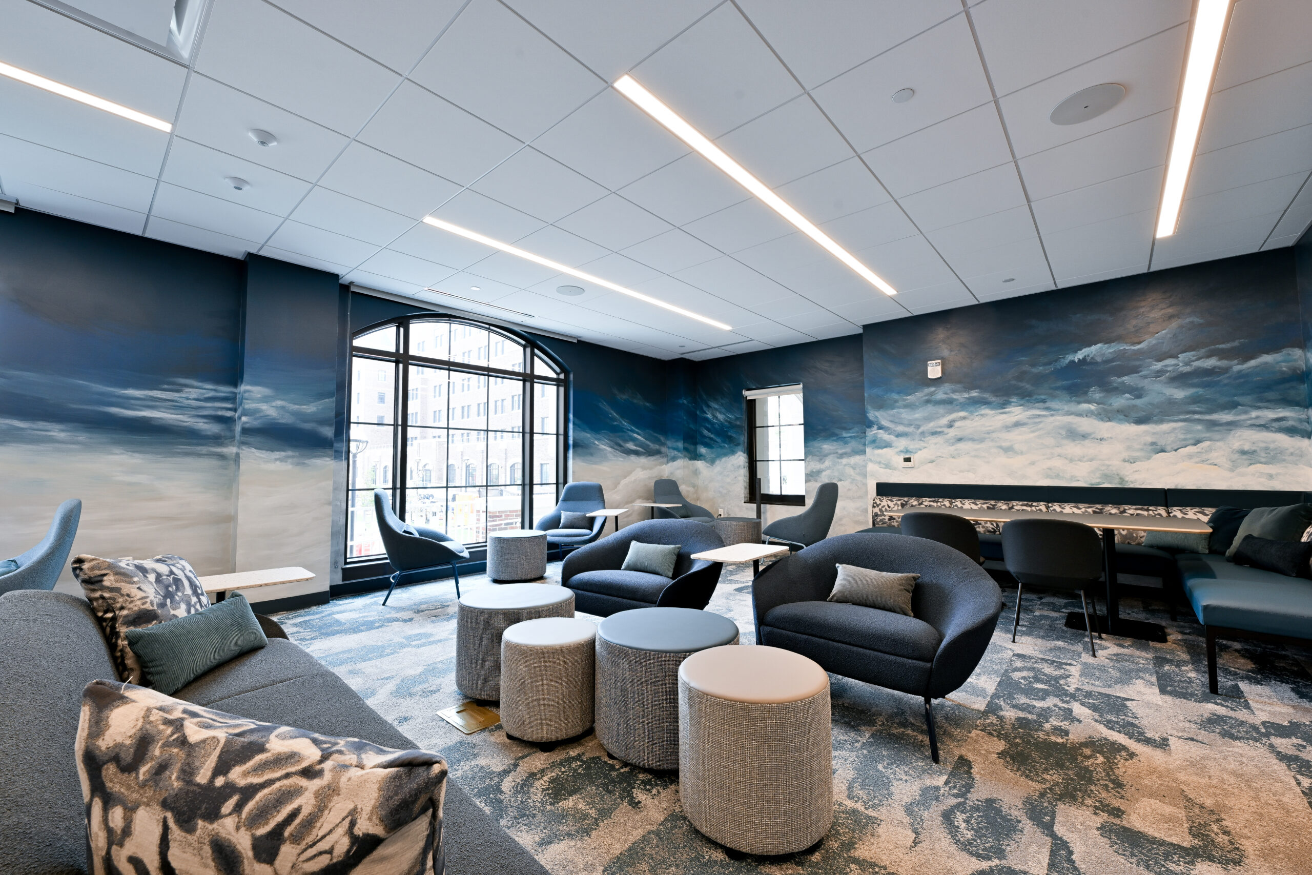 A modern lounge with dark blue walls featuring large cloud murals, contemporary seating, and a patterned carpet under bright ceiling lights.