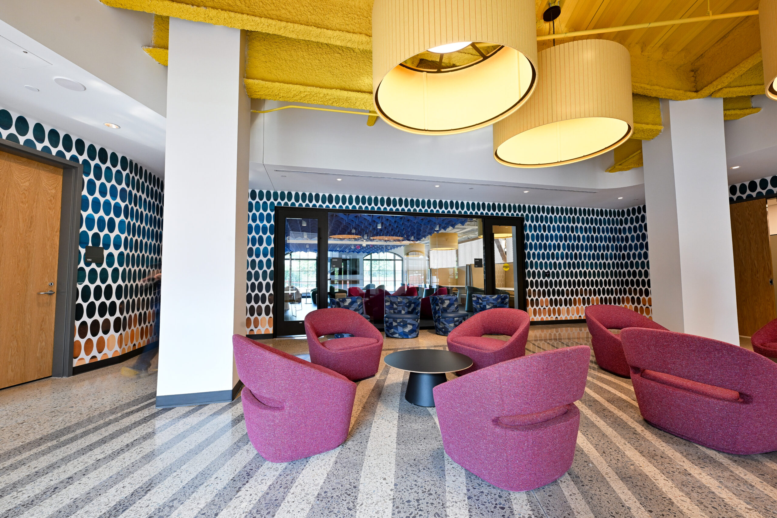 A modern lounge with vibrant pink armchairs, bold yellow ceiling beams, and patterned walls featuring circular designs.