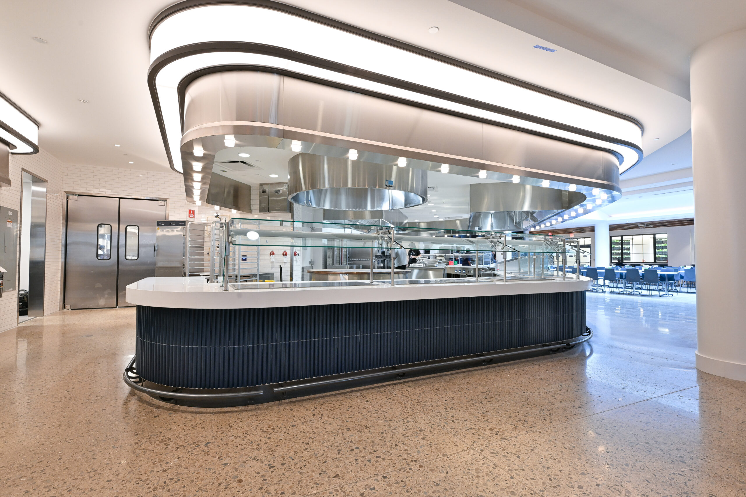 A modern, sleek food service counter with a curved design, stainless steel accents, and glass barriers, set in a bright, spacious dining area.