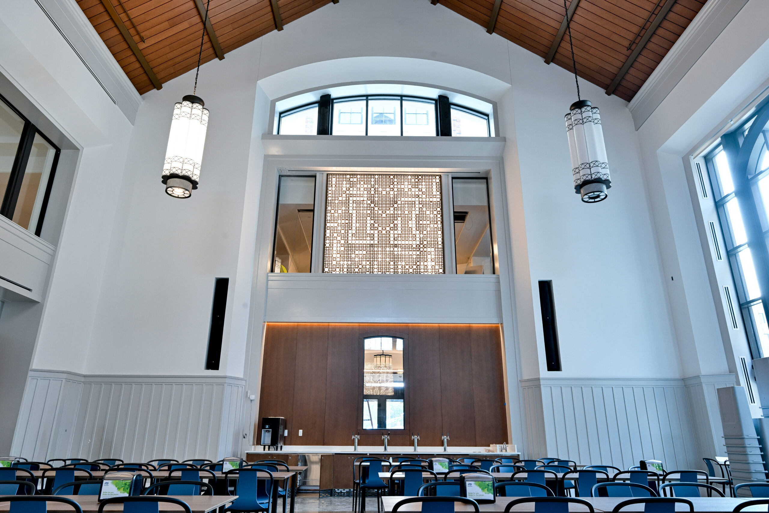 A modern, brightly lit hall with high ceilings, wooden beams, and rows of blue chairs arranged beneath hanging lanterns and large windows.