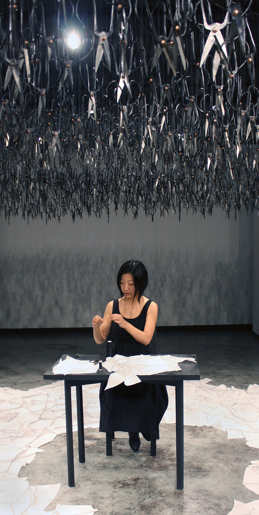 A person sits at a table with papers, under many scissors hanging from the ceiling in an art installation.