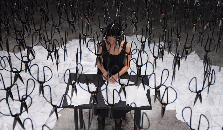 A person at a desk is surrounded by hanging scissors and white paper sheets scattered across the floor.