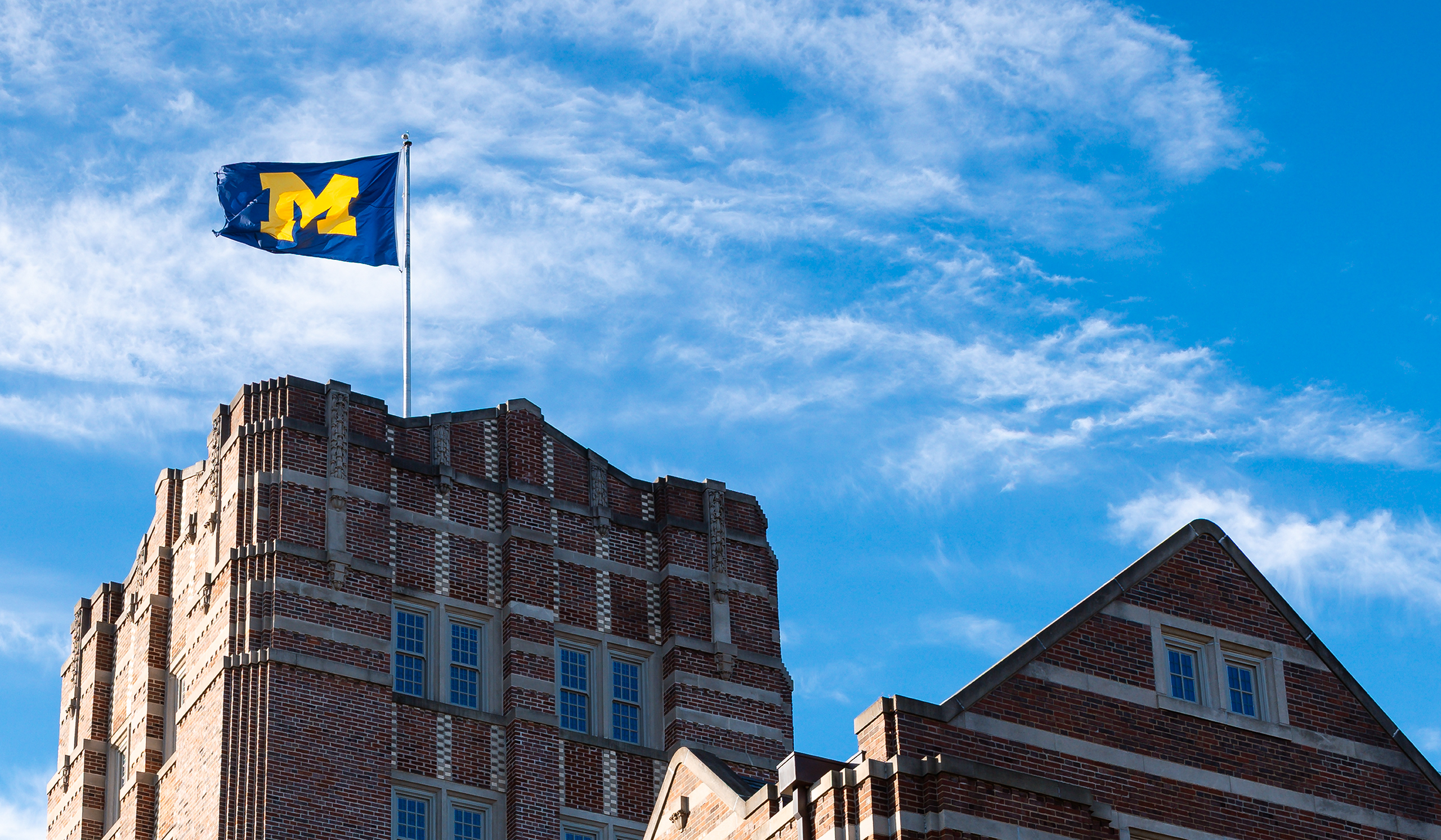 A blue Michigan flag, which features a maize Block M, flies atop the Michigan Union in front of a mostly blue sky.