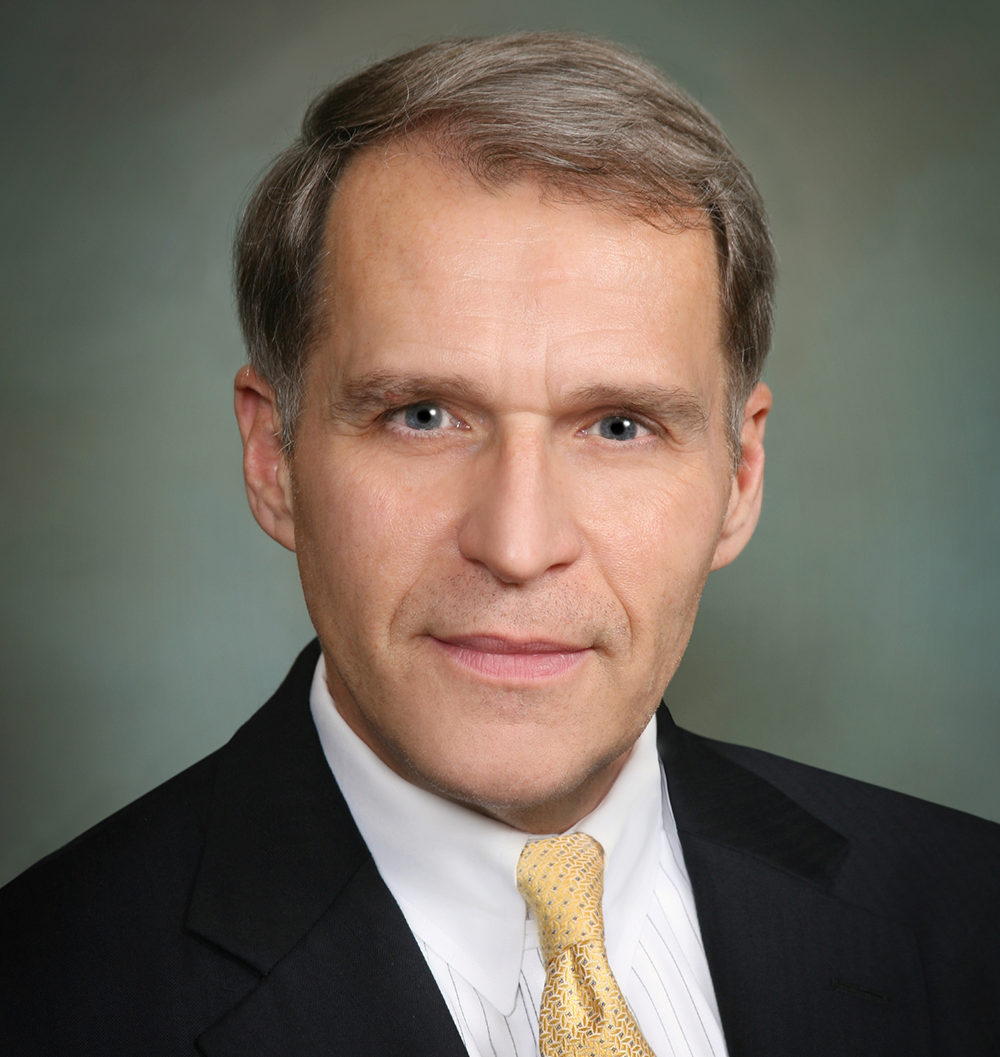 A person in a suit and yellow tie poses for a formal portrait against a plain, muted background.