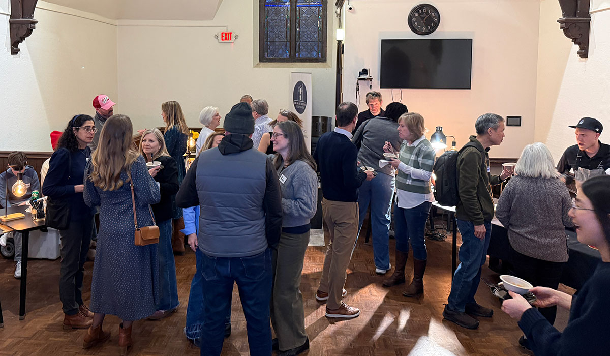 People gather to socialize and eat in a room with wooden floors, tables, and a wall-mounted TV.