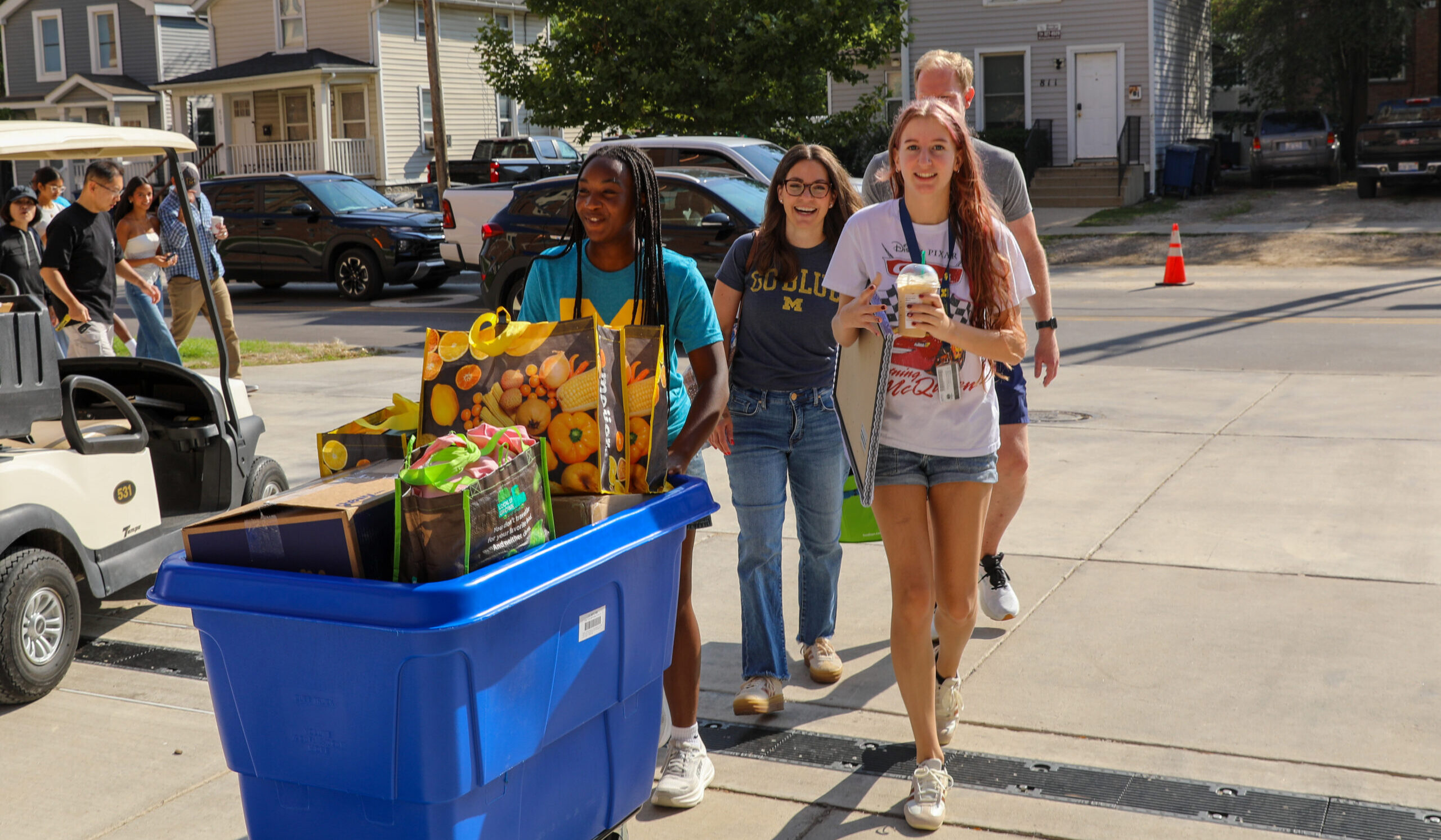 Three people push a blue bin filled with boxes and bags down a sidewalk on a sunny day.