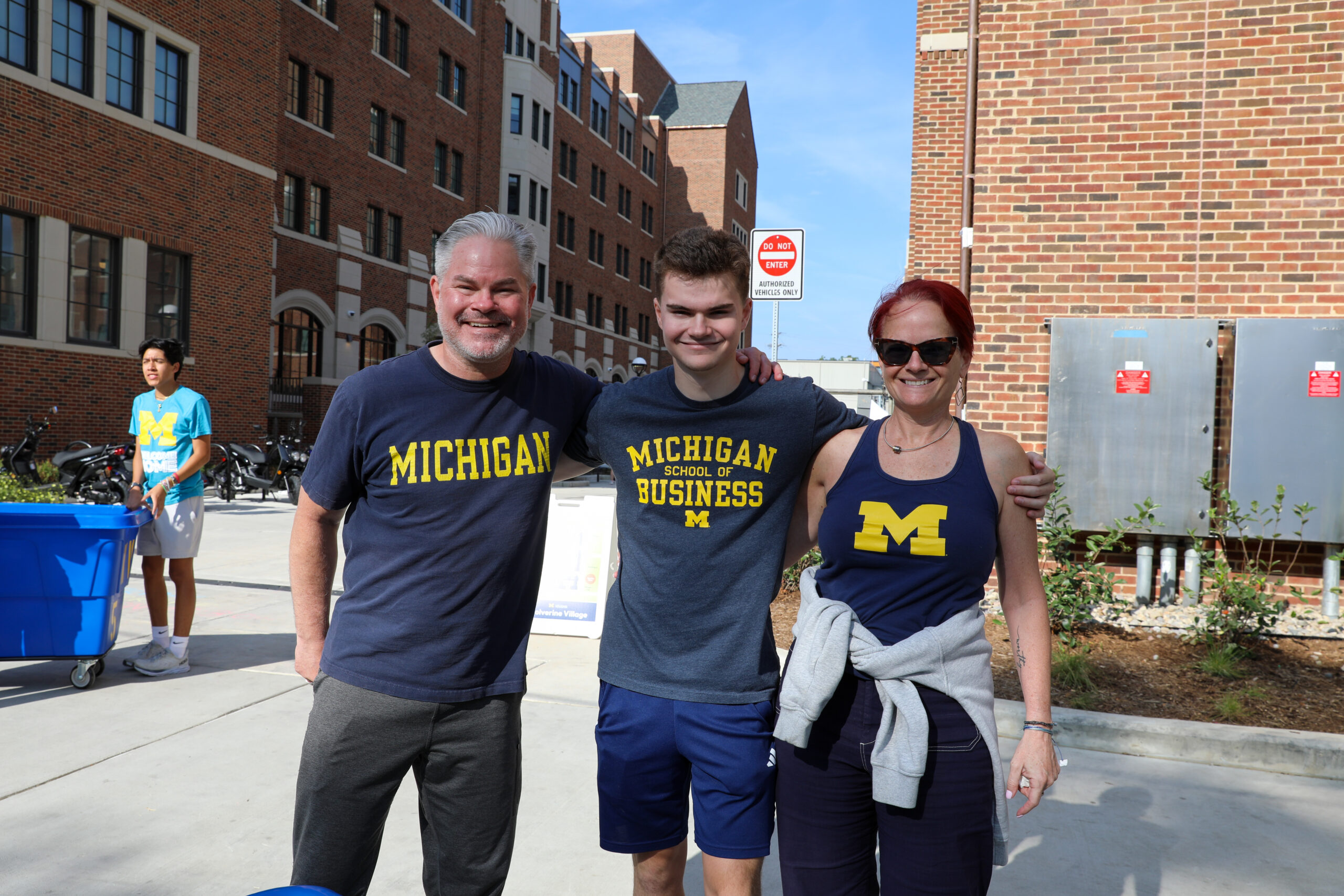A new student poses for a photo with his parents on move-in day. They are all wearing Michigan shirts.