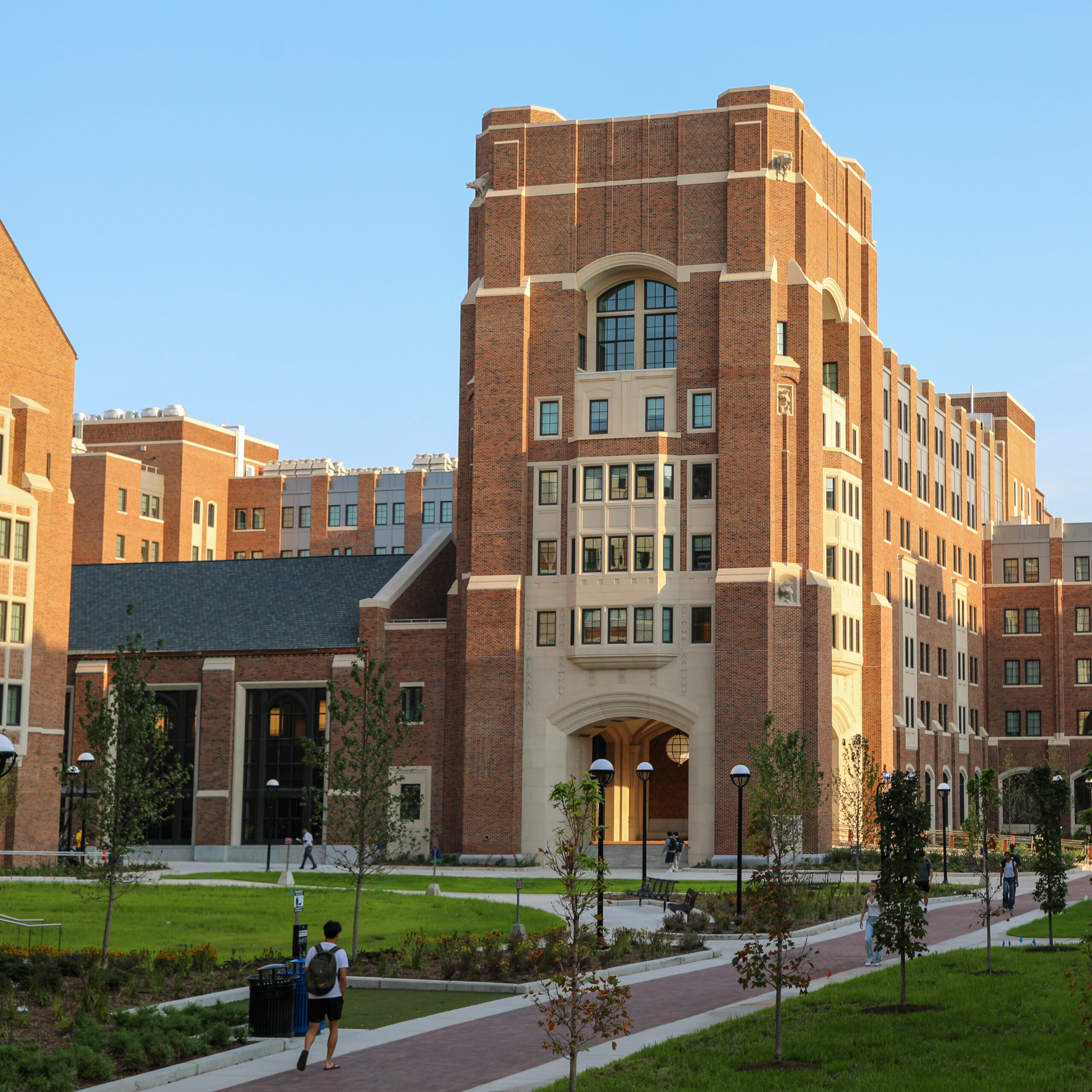 The brick tower of Wolverine Village, with blue skies and sun shining. Students walk in the courtyard, with brick pathways and green grass.