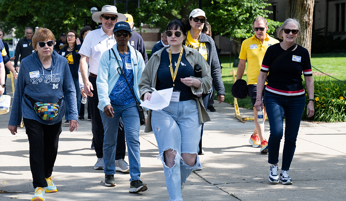 People wearing University of Michigan maize and blue gear walk together on a campus sidewalk.